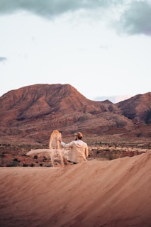 A person in casual clothing and a cap appears to be seated on a sand dune, sifting sand through their hand. The background features rugged, rocky mountains with a largely clear sky.