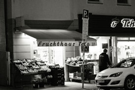 A black and white image featuring the exterior of a local fruit and vegetable shop named 'Fruchthaus Tela'. The storefront is illuminated, displaying various fruits and vegetables in crates along the entrance. A person is standing near the entrance, and a car is parked nearby. A signage board with the shop's name and house number is visible.
