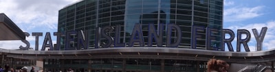 A large modern building facade with big blue letters spelling 'Staten Island Ferry'. The building features a glass exterior with tall, reflective windows, and a cloudy sky is visible in the background. Several people are walking in front of the structure.