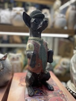 A statue of a dog dressed in human clothing, including a hat, vest, and tie, stands upright on its hind legs. The dog has a glossy finish and is detailed with realistic features. It is placed on a decorative base, and various ceramic objects are visible in the background, slightly out of focus.