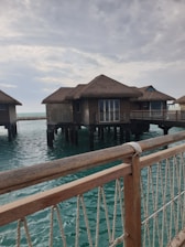 Several overwater bungalows with thatched roofs stand on stilts above a turquoise ocean. A wooden railing with ropes is visible in the foreground, and the sky is partly cloudy, casting a serene atmosphere over the scene.