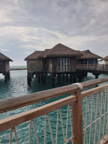 Several overwater bungalows with thatched roofs stand on stilts above a turquoise ocean. A wooden railing with ropes is visible in the foreground, and the sky is partly cloudy, casting a serene atmosphere over the scene.