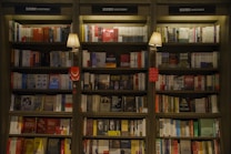 A well-organized bookshelf filled with a variety of books, mostly in Chinese, categorized under investment. The shelves are illuminated by warm light from two wall-mounted lamps, adding a cozy and inviting atmosphere to the space.