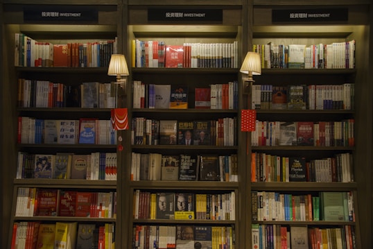 A well-organized bookshelf filled with a variety of books, mostly in Chinese, categorized under investment. The shelves are illuminated by warm light from two wall-mounted lamps, adding a cozy and inviting atmosphere to the space.