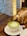 A cup of matcha latte with a heart-shaped design in the foam sits on a wooden table. A blurred background shows a light-colored brick wall and some wooden furnishings, including a pepper mill and napkin holder.