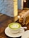 A cup of matcha latte with a heart-shaped design in the foam sits on a wooden table. A blurred background shows a light-colored brick wall and some wooden furnishings, including a pepper mill and napkin holder.