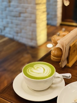 A cup of matcha latte with a heart-shaped design in the foam sits on a wooden table. A blurred background shows a light-colored brick wall and some wooden furnishings, including a pepper mill and napkin holder.