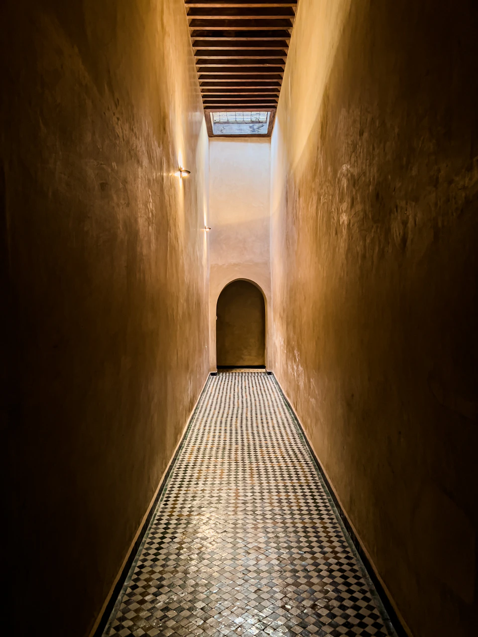 The image depicts a long, narrow hallway with a tiled floor and walls. The hallway is bathed in natural light streaming through an arched window on the right side, creating a warm glow that illuminates the space. On the left side of the hallway, there's a door marked by a small archway, adding architectural interest to the scene.

The tiled floor and walls are made of stone, giving the hallway a rustic charm. The image is taken from a low angle, making the hallway appear even more imposing as it stretches into the distance.