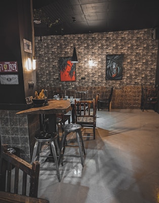 The image depicts a cozy, dimly lit interior of a restaurant or bar. It features a rustic wood table surrounded by assorted chairs and stools. The walls are adorned with artistic paintings, one displaying an abstract face in red and black tones. Overhead lighting adds a warm glow to the space, complementing the textured wall design.