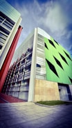 A modern architectural building with geometric design elements is prominently featured. The structure showcases a combination of glass and vibrant green panels with black triangular accents, alongside sections with red and beige details. The sky above is partly cloudy, adding a dramatic backdrop to the scene.