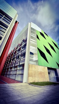 A modern architectural building with geometric design elements is prominently featured. The structure showcases a combination of glass and vibrant green panels with black triangular accents, alongside sections with red and beige details. The sky above is partly cloudy, adding a dramatic backdrop to the scene.
