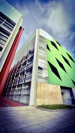 A modern architectural building with geometric design elements is prominently featured. The structure showcases a combination of glass and vibrant green panels with black triangular accents, alongside sections with red and beige details. The sky above is partly cloudy, adding a dramatic backdrop to the scene.