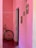 A bicycle with a rusted frame and white front wheel is leaning against a pink textured wall in a narrow hallway. The walls are adorned with small, framed decorations featuring natural elements like shells and stones. The floor appears to be made of dark, polished wood or laminate.