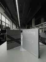 Two books are displayed on a table in an interior space with modern design elements. One book has a dark cover with striped shadows, while the other has a light cover with the title 'Building with History' and the author's name. The background reveals a spacious room with large windows, bookshelves, and contemporary lighting.