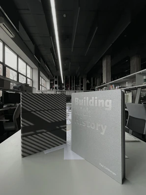 Two books are displayed on a table in an interior space with modern design elements. One book has a dark cover with striped shadows, while the other has a light cover with the title 'Building with History' and the author's name. The background reveals a spacious room with large windows, bookshelves, and contemporary lighting.