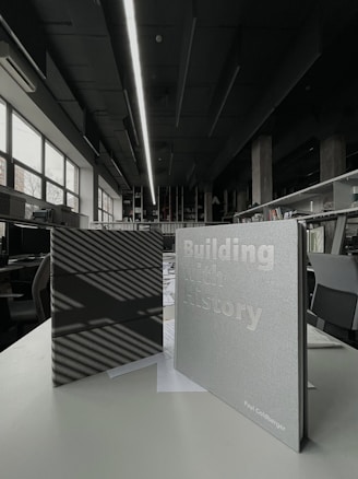 Two books are displayed on a table in an interior space with modern design elements. One book has a dark cover with striped shadows, while the other has a light cover with the title 'Building with History' and the author's name. The background reveals a spacious room with large windows, bookshelves, and contemporary lighting.