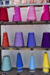 Multiple spools of yarn are arranged on a wooden shelf against a textured wall. The yarn is wrapped around conical spools in an array of vibrant colors including reds, pinks, purples, and blues.