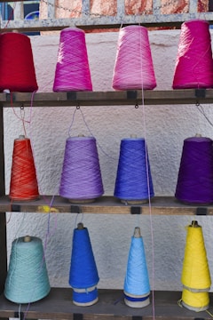 Multiple spools of yarn are arranged on a wooden shelf against a textured wall. The yarn is wrapped around conical spools in an array of vibrant colors including reds, pinks, purples, and blues.