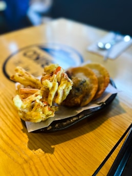A plate with several pieces of fried food, possibly tempura or fritters and another type of golden-brown fried item, placed on a sheet of parchment paper. The plate is situated on a light wooden table in a well-lit room. The background is slightly blurred, with what appears to be a fork and spoon resting on the table.