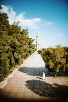 A long, paved walkway lined with lush green trees on both sides leads towards a tall, elegant tower or minaret in the distance. The clear blue sky contrasts with the greenery, while the shadows of the trees create interesting patterns on the path. A person dressed in white robes is walking along the path, adding a human element to the serene scene.