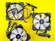 Three computer cooling fans are arranged on a bright yellow background. The fans have white blades with black frames. Each fan is accompanied by attached black cables.