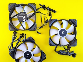 Three computer cooling fans are arranged on a bright yellow background. The fans have white blades with black frames. Each fan is accompanied by attached black cables.