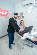 A dentist and a patient are in a dental office. The dentist, wearing scrubs, is pointing at a laptop on a mobile dental unit. The patient, seated in the dental chair, is looking at the screen with a smile. The room contains dental equipment and art featuring teeth on the wall.