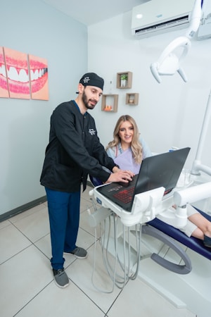 A dentist and a patient are in a dental office. The dentist, wearing scrubs, is pointing at a laptop on a mobile dental unit. The patient, seated in the dental chair, is looking at the screen with a smile. The room contains dental equipment and art featuring teeth on the wall.