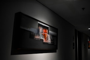 A modern, abstract painting is displayed on a dimly lit wall in what appears to be a hallway. The artwork features bold, geometric shapes with contrasting colors, primarily black, red, and white. The surrounding area is dark, enhancing the focus on the painting, which is partially illuminated by overhead lighting.