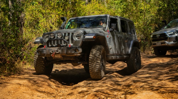A rugged off-road vehicle maneuvers through a dirt trail in a forested area. The vehicle has large tires and is coated with dust, indicating it has been driven through challenging terrain. Behind it, another vehicle is visible, partially covered by foliage.