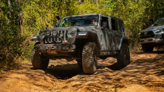 A rugged off-road vehicle maneuvers through a dirt trail in a forested area. The vehicle has large tires and is coated with dust, indicating it has been driven through challenging terrain. Behind it, another vehicle is visible, partially covered by foliage.