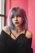 A person with pastel purple hair and dark, dramatic makeup gazes to the side. They are wearing a black off-shoulder top and a spiked necklace. The background consists of a vibrant pink wall, contributing a striking contrast to the overall look.