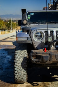 A rugged off-road vehicle with large, textured tires parked on a road. The vehicle's front grill and headlights are visible, along with additional lights and equipment on the hood and roof. The background shows a rural landscape with a clear sky and distant mountains.