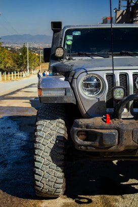 A rugged off-road vehicle with large, textured tires parked on a road. The vehicle's front grill and headlights are visible, along with additional lights and equipment on the hood and roof. The background shows a rural landscape with a clear sky and distant mountains.