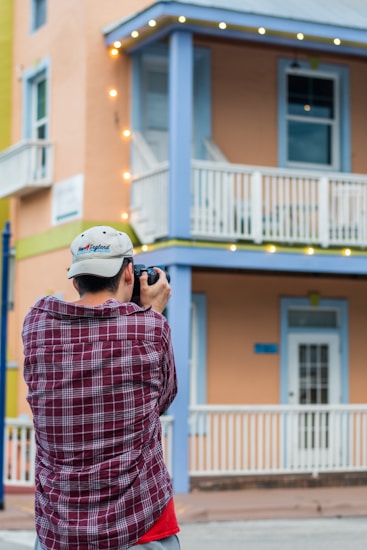 A person wearing a plaid shirt and a cap is taking a photo of a two-story building with pastel-colored walls. The building features a balcony with white railings and is adorned with string lights.