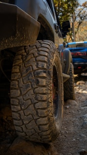 A close-up view of a rugged off-road vehicle's tire, caked with dirt and mud, set against a rocky terrain. In the background, another vehicle can be seen, slightly blurred and also positioned on what appears to be a challenging off-road path. The surrounding foliage hints at a natural, wooded environment.