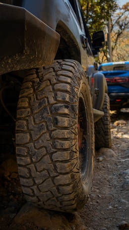 A close-up view of a rugged off-road vehicle's tire, caked with dirt and mud, set against a rocky terrain. In the background, another vehicle can be seen, slightly blurred and also positioned on what appears to be a challenging off-road path. The surrounding foliage hints at a natural, wooded environment.