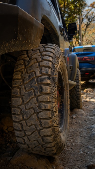 A close-up view of a rugged off-road vehicle's tire, caked with dirt and mud, set against a rocky terrain. In the background, another vehicle can be seen, slightly blurred and also positioned on what appears to be a challenging off-road path. The surrounding foliage hints at a natural, wooded environment.
