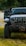 The front view of an off-road vehicle, likely a Jeep, with large tires and a winch attached to the front bumper. The vehicle is parked on grass in a wooded area with another car visible in the background. The driver's door is open.