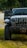 The front view of an off-road vehicle, likely a Jeep, with large tires and a winch attached to the front bumper. The vehicle is parked on grass in a wooded area with another car visible in the background. The driver's door is open.