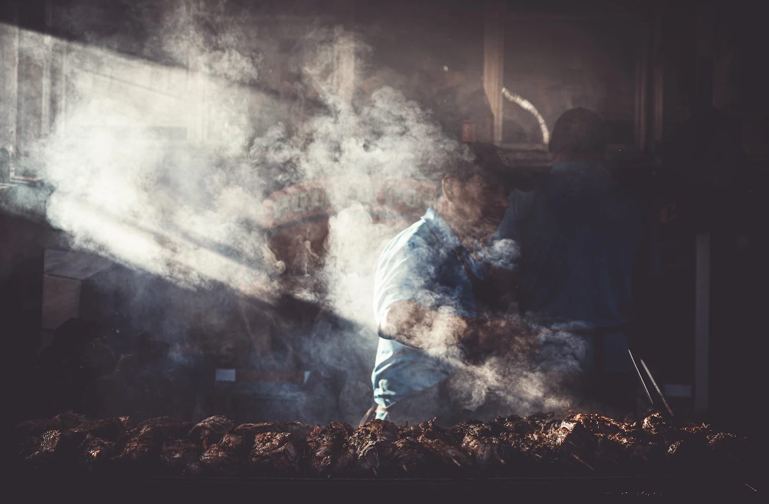 A pitmaster half-hidden in a column of wood smoke above the long smoker grate