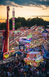 A vibrant amusement park bustling with people enjoying various rides and attractions under a twilight sky. Brightly lit signs advertise games and food stalls, including funnel cakes and corn dogs. Roller coasters and thrill rides are prominent, with colorful lights illuminating the scene.