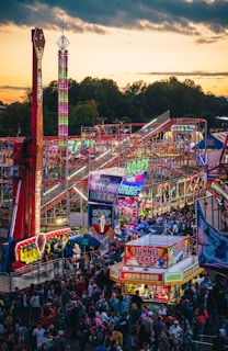 A vibrant amusement park bustling with people enjoying various rides and attractions under a twilight sky. Brightly lit signs advertise games and food stalls, including funnel cakes and corn dogs. Roller coasters and thrill rides are prominent, with colorful lights illuminating the scene.