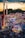 A vibrant amusement park bustling with people enjoying various rides and attractions under a twilight sky. Brightly lit signs advertise games and food stalls, including funnel cakes and corn dogs. Roller coasters and thrill rides are prominent, with colorful lights illuminating the scene.