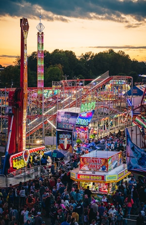 A vibrant amusement park bustling with people enjoying various rides and attractions under a twilight sky. Brightly lit signs advertise games and food stalls, including funnel cakes and corn dogs. Roller coasters and thrill rides are prominent, with colorful lights illuminating the scene.