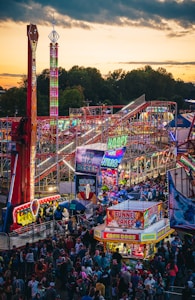 A vibrant amusement park bustling with people enjoying various rides and attractions under a twilight sky. Brightly lit signs advertise games and food stalls, including funnel cakes and corn dogs. Roller coasters and thrill rides are prominent, with colorful lights illuminating the scene.