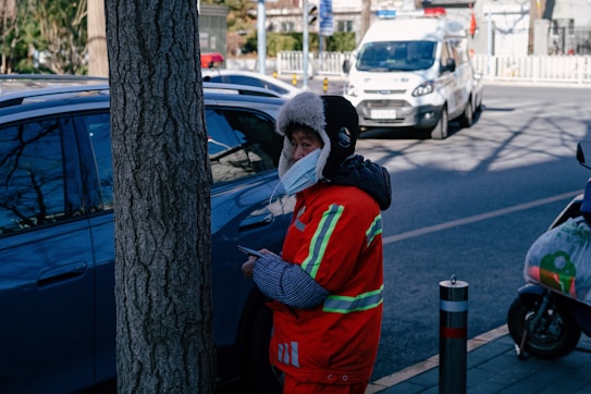 A person wearing a bright orange reflective jacket and a furry hat is standing by a tree on a city street. They are wearing a face mask and appear to be holding a small object in their hands. In the background, there are parked vehicles including a van and a scooter, with urban buildings visible in the distance.