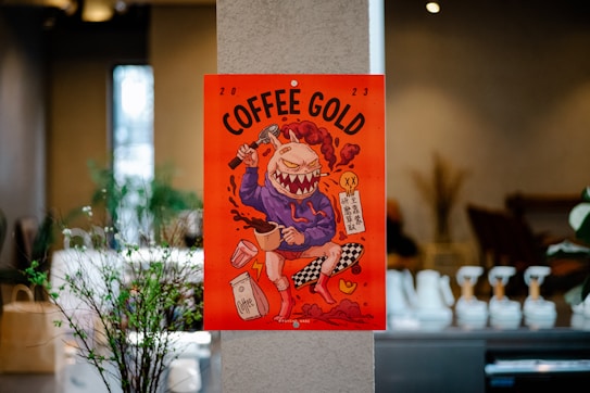 A colorful and vibrant poster featuring a cartoon-like character with exaggerated features, including large, sharp teeth and fiery hair. The character appears to be holding a coffee cup and a hammer, with coffee bags and splashes surrounding it. The poster has bold text saying 'COFFEE GOLD' and is placed against a neutral background in an indoor setting.