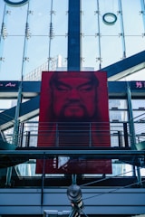 A large, red portrait is displayed on a modern building with glass panels and metal structures. The architectural elements create a geometric pattern, and the portrait features a face with a stern expression. The glass background reflects parts of the city skyline, adding depth to the composition.