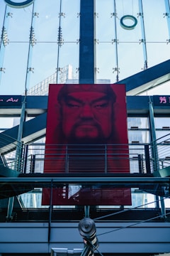 A large, red portrait is displayed on a modern building with glass panels and metal structures. The architectural elements create a geometric pattern, and the portrait features a face with a stern expression. The glass background reflects parts of the city skyline, adding depth to the composition.
